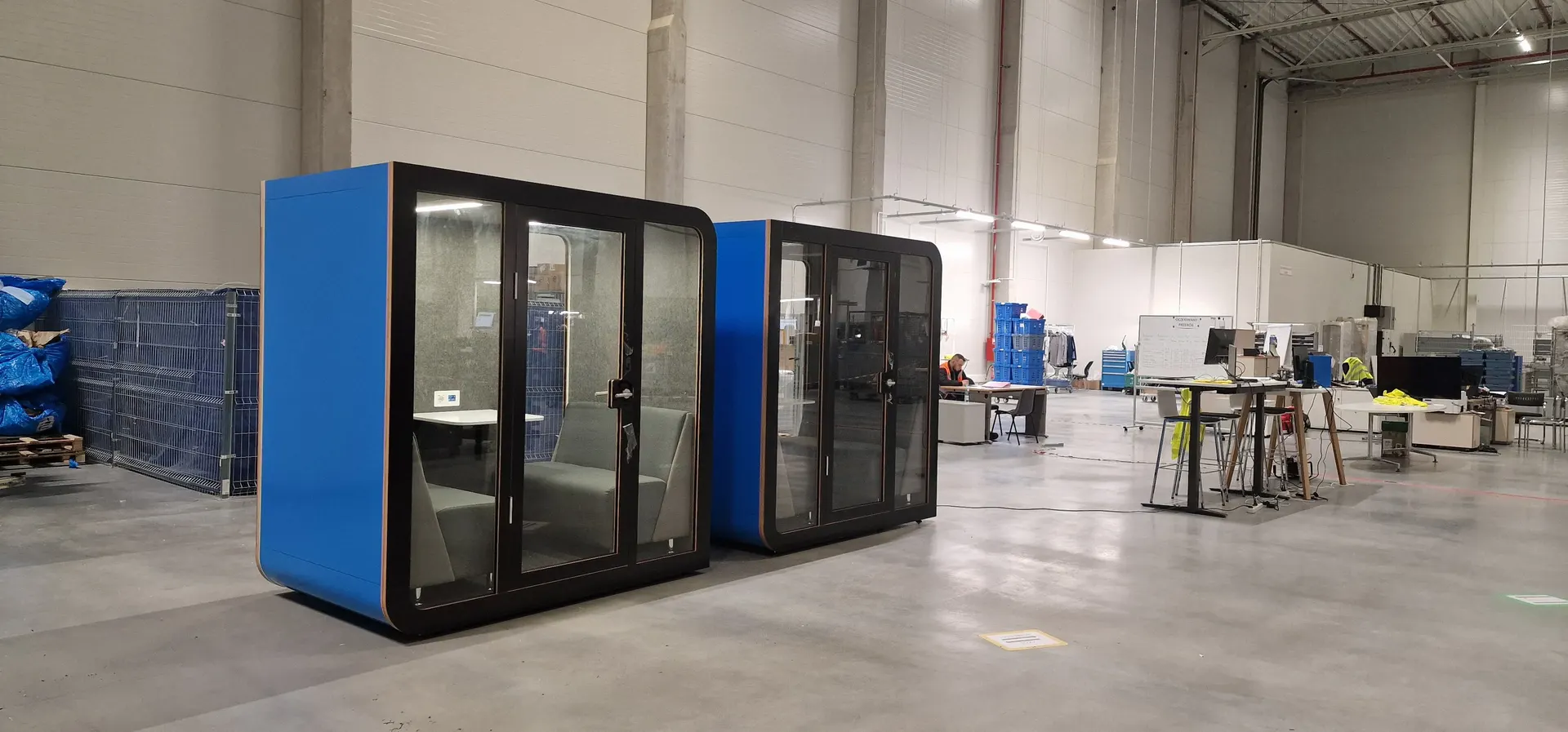 Two blue Silentbox Duet acoustic booths with dark frames standing on the floor of a logistics warehouse, workbenches and staff in the background.