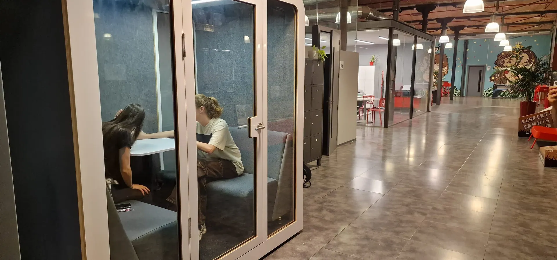 Two people sitting opposite each other inside a white Silentbox Duet booth in a coworking space, seen through the glass door.
