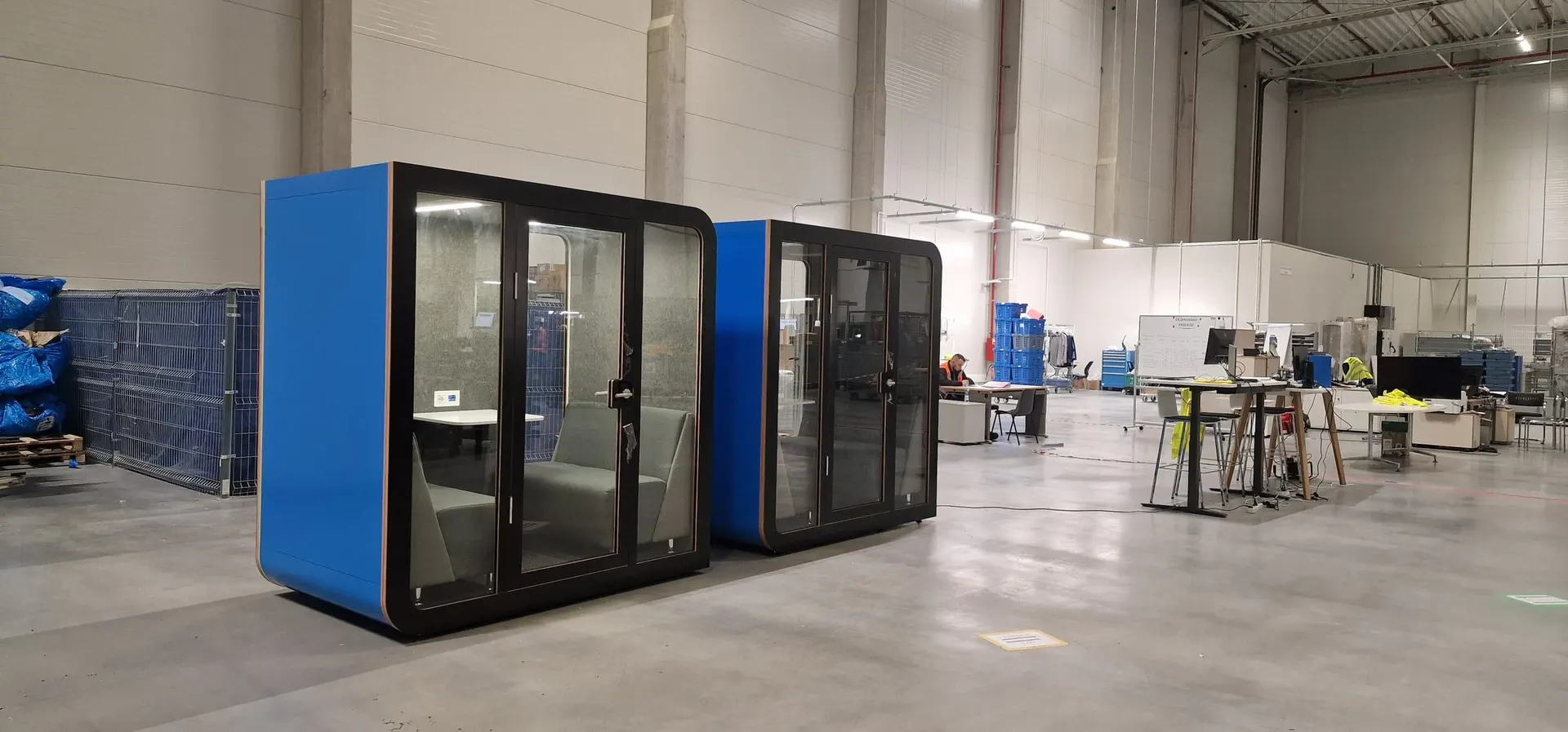 Two blue Silentbox Duet acoustic booths with dark frames standing on the floor of a logistics warehouse, workbenches and staff in the background.