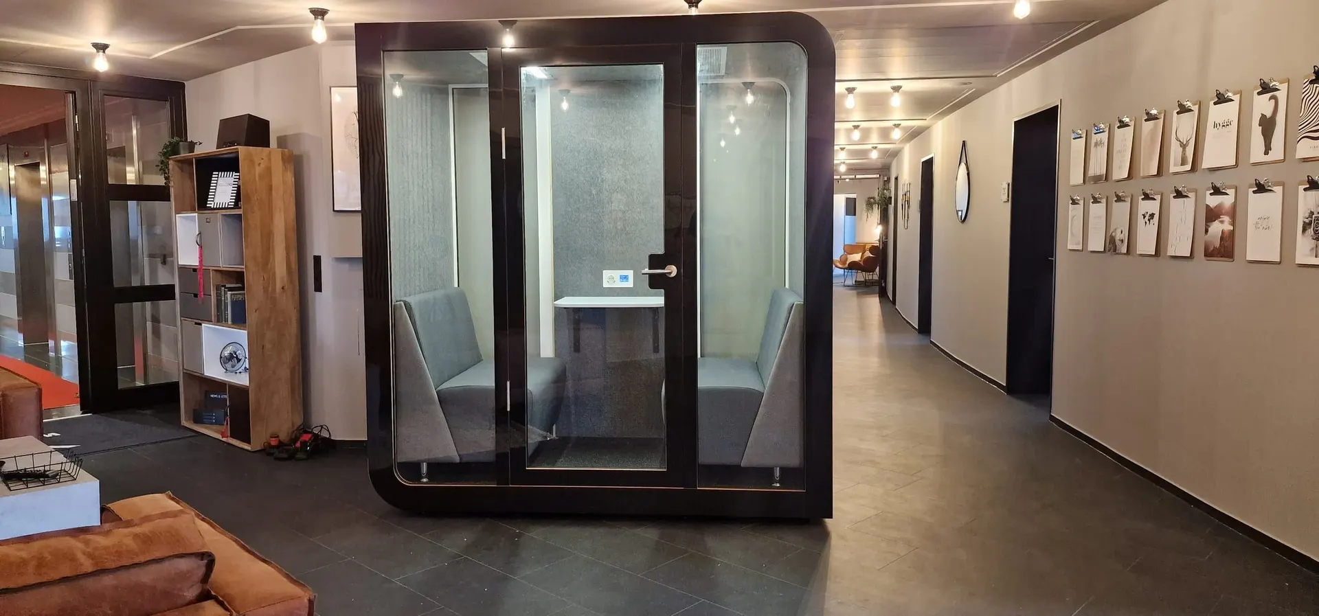 A black four-person Silentbox Quartet booth with two facing bench seats and a table, standing in an office corridor lined with framed prints.